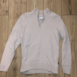 Beige tröja med dragkedja från Casual Friday - Säljer en stilren beige tröja från Casual Friday. Tröjan har en dragkedja vid halsen och är tillverkad i ett texturerat material. Perfekt för en avslappnad look. Har aldrig använts 