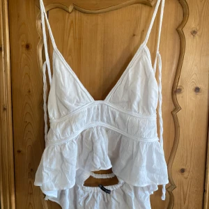 Vit ärmlös topp från Zara - Säljer en vit ärmlös topp från Zara med smala axelband och volangdetaljer. Toppen har en luftig och somrig design med en v-ringad framdel. Perfekt för varma dagar!