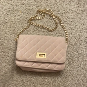 Rosa quiltad axelväska med guldkedja - Snygg beige axelväska med quiltat mönster och guldkedja som axelrem. Väskan har en elegant låsdetalj i guld på framsidan och en praktisk innerficka med dragkedja. Perfekt för att ge en touch av lyx till din outfit.