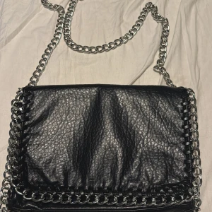 Svart axelväska från Tiamo - Snygg svart axelväska från Tiamo i skinnimitation med kedjedetaljer. Väskan har en dragkedja på baksidan och en kedja som axelrem. Perfekt för att ge din outfit en edgy touch.