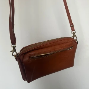 Brun axelväska i skinn - Snygg brun axelväska i skinn med justerbar rem och dragkedja. Perfekt för att bära dina nödvändigheter med stil. Enkel och elegant design som passar till många tillfällen. Dragkedjan på lilla facket är tyvärr borta.
