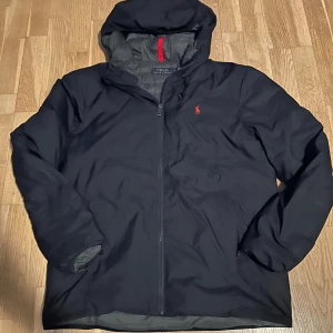 Mörkblå jacka från Ralph Lauren - Snygg mörkblå jacka från Ralph Lauren med huva och dragkedja. Jackan har en diskret röd logga på bröstet och är perfekt för kyligare dagar. Den har en quiltad insida för extra värme och komfort.