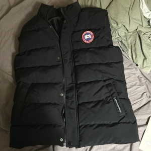 Svart dunväst från Canada Goose - Säljer en svart dunväst från Canada Goose med deras ikoniska logga på bröstet. Västen har en dragkedja och knappar framtill samt två fickor. Perfekt för kyliga dagar när du vill hålla värmen utan att kompromissa med stilen.