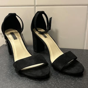 Svarta klackskor från Nelly - Snygga svarta klackskor från Nelly i ”mocka” med öppen tå och justerbart spänne runt ankeln. Perfekta för en stilren look. Använd endast vid två korta tillfällen. Kom gärna med ett prisförslag om du är intresserad🖤