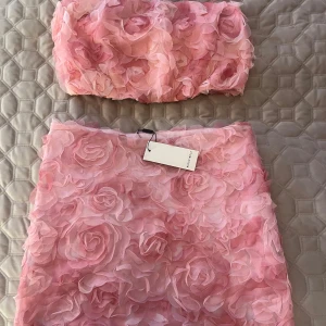 Rosa set med blommor från Rut&Circle - Säljer ett superfint rosa set med kjol och bandeutopp från Rut&Circle med 3D-blommor över hela tyget. Kjolen har en kort längd och är perfekt för att sticka ut med sin unika design. Matcha med den medföljande tubtoppen för en komplett look! Aldrig använt och helt nytt med prislappen kvar. Priset är för hela settet💕