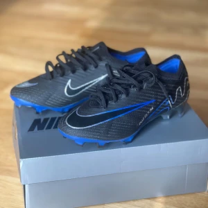 Nike Air Zoom Mercurial fotbollsskor - Snygga Nike Air Zoom Mercurial fotbollsskor i svart och blått. Skorna har en modern design med snörning och är perfekta för fotbollsplanen. De har en slitstark sula och är gjorda för att ge bra grepp och snabbhet.