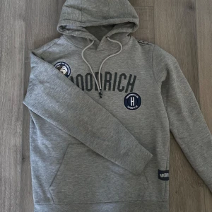Grå hoodie från Hoodrich - Säljer en snygg grå hoodie från Hoodrich med tryck och broderade detaljer på framsidan. Den har en justerbar huva med dragsko och långa ärmar. Perfekt för en avslappnad stil. St S