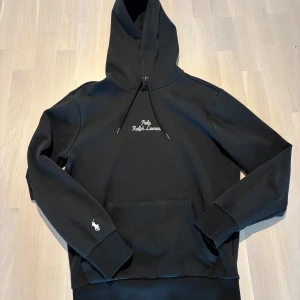 Svart hoodie från Ralph Lauren - Snygg svart hoodie från Ralph Lauren med broderad logga på bröstet och en liten logga på ärmen. Den har en klassisk känguruficka och justerbar huva med snören. Perfekt för en stilren och bekväm look.