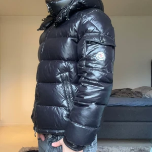 Svart Maya från Moncler - Snygg svart dunjacka från Moncler i storlek 1 med glansig finish och dragkedja framtill. Jackan har en hög krage och en logotyp på ärmen. Perfekt för kyliga dagar. Säljer då vinterdagarna börjar ta slut. För fler frågor eller bilder, kontakta.