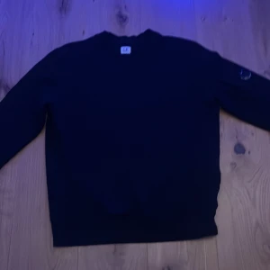 Svart tröja från C.P. Company - Säljer en stilren svart tröja från C.P. Company. Tröjan har en rund hals och en unik detalj med en lins på ärmen. Perfekt för en minimalistisk look.
