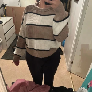 Randig stickad tröja i beige och vit - Mysig stickad tröja med breda ränder i beige, vit och svart. Tröjan har en avslappnad passform och långa ärmar, perfekt för kyligare dagar. Den ribbade halsringningen ger en klassisk look. Den passar dig som har storlek m/s