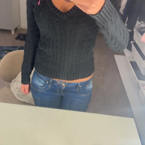 Mörkgrå kabelstickad tröja från Ralph Lauren - Säljer en snygg mörkgrå kabelstickad tröja från Ralph Lauren med en rosa logga på bröstet. Tröjan har en v-ringad design och långa ärmar. Perfekt för en stilren look!