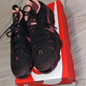 Nike Air Vapormax 2023 FK - Snygga Nike Air Vapormax 2023 FK i svart och vinröd färg. Skorna har en stickad ovandel med snörning och en unik sula i vinrött. Perfekta för en sportig look med komfort och stil. De är endast testade och därmed i princip helt nya! Var lite för små för mig och bara haft dem på hyllan. Köpta på JD
