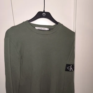 Grön långärmad tröja från Calvin Klein Jeans - Säljer en stilren grön långärmad tröja från Calvin Klein Jeans. Tröjan har en rund halsringning och ett märke med CK-logga på ärmen. Perfekt för en avslappnad look. Passar M 