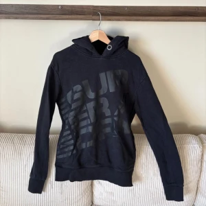Svart hoodie från Superdry - Säljer en svart hoodie från Superdry med stort tryck på framsidan. Hoodien har en justerbar huva med metallögla och ribbade muddar vid ärmslut och nederkant. Perfekt för en avslappnad stil.