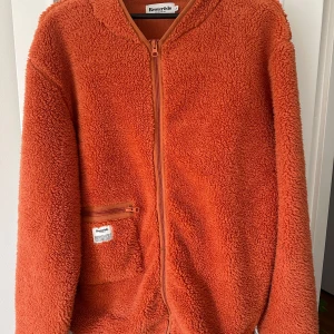 Orange teddyjacka från Resteröds - Säljer en mysig orange teddyjacka från Resteröds. Jackan har en dragkedja framtill och en praktisk ficka med dragkedja på sidan. Köpt men aldrig använd. 