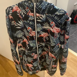 Mönstrad jacka med flamingos från Blount & Pool - Säljer en cool jacka från Blount & Pool med ett tropiskt mönster av flamingos och blad. Jackan har en dragkedja framtill och är långärmad. Perfekt för att sticka ut med en unik stil!