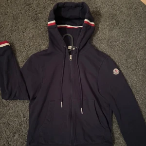 Mörkblå hoodie från Moncler - Pris kan diskuteras kom PM‼️                    Snygg mörkblå hoodie från Moncler med dragkedja och huva. Den har röda och vita ränder på huvan och ärmsluten. Perfekt för en stilren och bekväm look.                                         Storlek S men passar även XS‼️