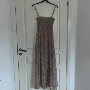 Blommig långklänning från SNOB - Söt långklänning från SNOB med smala axelband och smockad överdel. Axelbanden går att ta loss. Klänningen har ett blommigt mönster i rosa, grönt och vitt och är tillverkad i 100% bomull. Perfekt för varma dagar och en romantisk look.