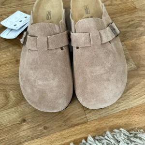  Birkenstock boston tofflor i mocka - Säljer ett par klassiska beige Birkenstock tofflor i mjuk mocka med justerbart spänne på sidan. Skorna har en platt sula och rund tå, perfekt för en avslappnad stil. Ikonisk komfort och tidlös design. Säljer för torde de passa så slängde kartongen! Men de visa sig va för små! Så skulle säga att de passar dig som har 39,5