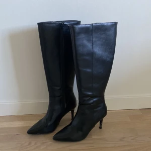 Svarta höga stövlar med klack - Säljer ett par svarta höga stövlar i skinnimitation med spetsig tå och smal klack. Stövlarna har dragkedja på insidan och en elegant, stilren design som passar till många outfits.