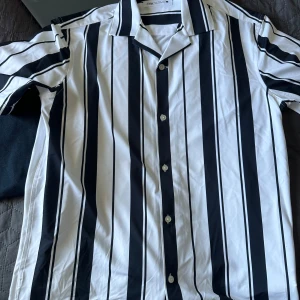 Vit och svart randig skjorta från Zara - Säljer en snygg kortärmad skjorta från Zara med breda svarta och smala vita ränder. Skjortan har klassisk krage och knäppning framtill. Perfekt för dig som gillar stilrena mönster och vill sticka ut lite extra.