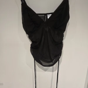 Svart mesh linnetopp från H&M Divided - Säljer en svart mesh linnetopp från H&M Divided. Toppen har smala axelband, v-ringning och är croppad med snörning framtill. Perfekt för dig som gillar trendiga och lätta plagg med en edgy vibe.