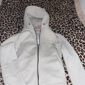 Vit hoodie från Moncler - Säljer en stilren vit hoodie från Moncler med dragkedja framtill och huva. Tröjan har långa ärmar och två fickor på sidorna. Perfekt för en avslappnad och trendig look.