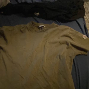 Olivgrön t-shirt från Moncler - Säljer en olivgrön t-shirt från Moncler med tryckt logga vid halsen och ett broderat Moncler-märke på ärmen. Klassisk passform och rund halsringning. Perfekt för dig som gillar stilrena märkesplagg.