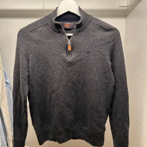 Mörkgrå merinoullströja med dragkedja från Morris - Snygg mörkgrå tröja från Morris i 100% merinoull. Tröjan har halv dragkedja framtill och ribbade muddar vid ärmslut och nederkant. Perfekt för lager-på-lager och stilren look. Tröjan är krympt från M till XS och är i väldigt bra skick