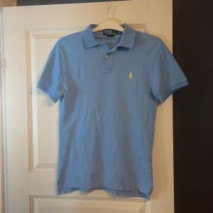 Ljusblå pikétröja från Polo Ralph Lauren - Säljer en snygg ljusblå pikétröja från Polo Ralph Lauren i storlek S. Tröjan har en klassisk krage med knappar och det ikoniska broderade logotypen i gult på bröstet. Perfekt för en stilren och avslappnad look!