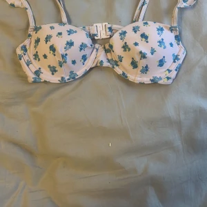 Blommig bikiniöverdel - Söt bikiniöverdel i vitt med blått blommönster från good collection. Har bygel och justerbara axelband samt spänne framtill. Tillverkad i återvunnen polyester.
