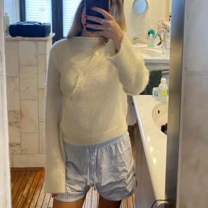 Vit stickad tröja med sned knäppning - Mysig vit stickad tröja med lång ärm och unik sned knäppning framtill. Tröjan har en ribbad kant vid hals, ärmslut och nederkant. Perfekt för kyligare dagar och enkel att matcha med olika outfits.