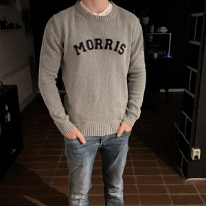 Grå stickad tröja från Morris - Säljer en stilren grå stickad tröja från Morris med svart broderad logga på bröstet. Tröjan har rund halsringning och ribbade muddar vid ärmslut och nederkant. Perfekt för lager-på-lager och enkel att matcha.