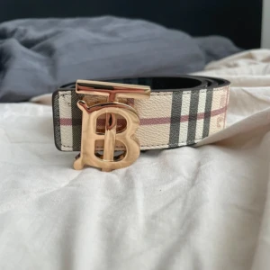 Burberry bälte med guldspänne - Säljer ett Burberry bälte med klassiskt rutigt mönster och stort guldigt TB-spänne. Ena sidan är beige med Burberrys signaturrutor, andra sidan är svart med diskret rutmönster och logga. Bältet är i skinn och passar perfekt till att lyfta din outfit.