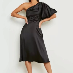 Svart satinklänning med one shoulder - Snygg svart klänning från Boohoo i satin med en axel och puff ärm i storlek 36. Klänningen har en elegant omlottkänsla vid midjan och faller till under knäet. Perfekt för dig som vill sticka ut med en unik siluett och lyxig känsla.