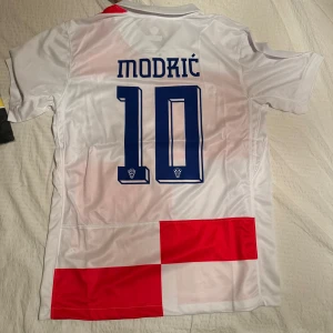 Kroatiens matchtröja från Nike MODRIC - Säljer en vit och röd fotbollströja från Nike med nummer 10 och namn på baksidan. Tröjan har korta ärmar och ett rutigt mönster i rött och vitt. Perfekt för fotbollsfans som vill visa sitt stöd! 🏆(prislapp sitter kvar)