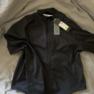 Svart träningsjacka från 157 - Svart träningsjacka från 157 med dragkedja framtill och hög krage. Jackan är tillverkad i ett lätt syntetmaterial och har en sportig look, perfekt för träning eller aktiva dagar. En lululemon liknande jacka!