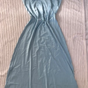 Ljusblå långklänning med smala axelband - Säljer en elegant ljusblå långklänning i satin med smala axelband och knytning i ryggen. Klänningen har en mjuk, glansig yta och figurnära passform som ger en stilren look. Perfekt för sommarens speciella tillfällen.