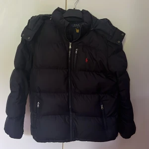 Marinblå dunjacka från Polo Ralph Lauren - Säljer en svart dunjacka från Polo Ralph Lauren med huva och dragkedja framtill. Jackan har två fickor med dragkedja och den klassiska röda loggan broderad på bröstet. Perfekt för kyliga dagar. Finns väldigt små defekter på den.