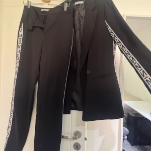 Svart kostym med vita loggband från NA-KD - Svart kostym från NA-KD med raka byxor och kavaj. Både byxorna och kavajen har vita band med svart logotyp längs sidorna. Kavajen har två knappar och fickor framtill. 