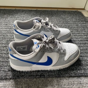 Nike Dunk Low i grått och vitt med blå swoosh - Nike Dunk Low sneakers i grått och vitt med en blå swoosh på sidan. Använt få gånger, skorna har snörning, rund tå och platt sula. Perfekta för dig som gillar klassisk streetstil och vill ha något som sticker ut lite extra. Extremt bra kvalité och inga defekter! För ENDAST 700kr så får du de snyggaste skor! Pris kan diskuteras. I storlek 37,5! 