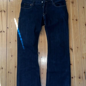 Mörkblå bootcut jeans raw denim - Snygga mörkblå jeans med bootcut passform och klassiska fickor fram och bak. Jeansen har kontrastsömmar och stängs med knapp och dragkedja. Perfekta för dig som gillar en tidlös stil med lite vidare ben.
