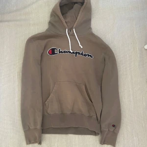 Beige hoodie från Champion - Säljer en beige hoodie från Champion med stor broderad logga på bröstet och klassisk känguruficka. Tröjan har huva med vita snören och ribbade muddar. Perfekt för en avslappnad stil. Den har många små skavanker.