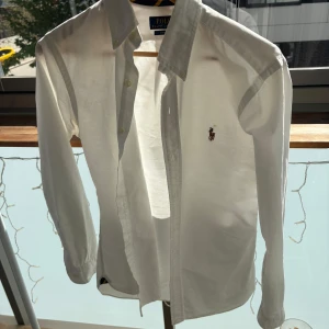 Vit skjorta från Polo Ralph Lauren - Klassisk vit skjorta från Polo Ralph Lauren i slim fit-modell. Skjortan har långa ärmar, knappar framtill och den ikoniska broderade loggan på bröstet. Perfekt för en stilren och tidlös look. Säljer för den har blivit för liten så passformen på mig är inte så bra kan ni tänka på!