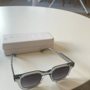 Grey 04 solglasögon från CHIMI - Snygga solglasögon från CHIMI med transparent grå båge och fyrkantig form. Glasen är mörktonade och bågarna är i plast. Perfekta för att lyfta din stil med en modern touch. Medföljer originaletui och putsduk.