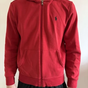 Röd hoodie från Ralph Lauren - Säljer en klassisk röd hoodie från Ralph Lauren med dragkedja och två fickor framtill. Tröjan har en liten broderad logga på bröstet och huva. Perfekt för en avslappnad stil.