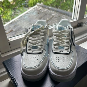 Nike Air Force 1 Shadow Hydrogen Blue - Nike Air Force 1 Shadow i vitt med detaljer i ljusblått och svart. Skorna har klassisk snörning, perforerad tå och en chunky sula. Swoosh-logga i svart på sidan och blå Nike-detaljer på plösen och hälen. Perfekta sneakers för dig som gillar stilrena men unika detaljer. Helt nya (oanvända).
