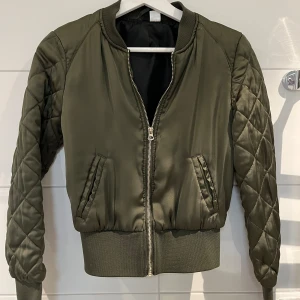 Olivgrön bomberjacka med quiltade ärmar - Snygg olivgrön bomberjacka med quiltade långärmade ärmar och breda muddar. Jackan har dragkedja framtill och två fickor med dekorativa detaljer. Perfekt för dig som vill ha en stilren och trendig look.