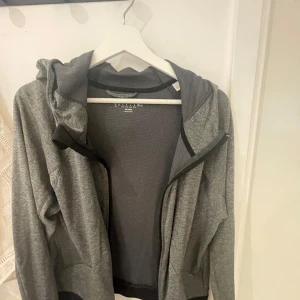 Grå hoodie med dragkedja från adidas - Sportig grå hoodie från adidas med hel dragkedja och huva. Jackan har svarta muddar och fickor framtill. Perfekt för träning eller en avslappnad stil. Tillverkad i mjukt syntetmaterial för extra komfort.